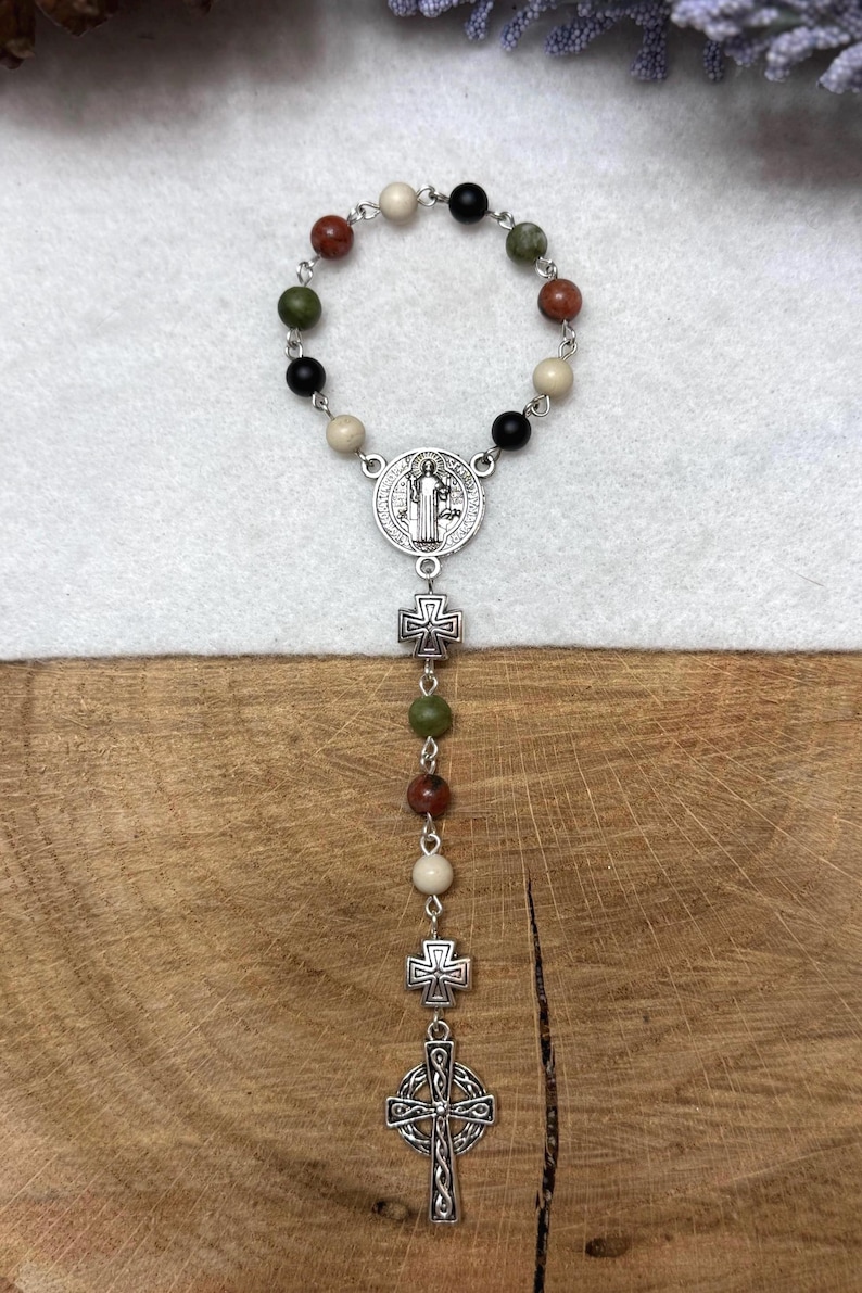 Puede incluir: Un rosario de una d&eacute;cada con una disposici&oacute;n circular de cuentas de piedra multicolor en tonos negro, blanco, verde y marr&oacute;n rojizo. Incluye una medalla de San Benito plateada, dos peque&ntilde;os espaciadores en forma de cruz y un colgante de cruz celta.