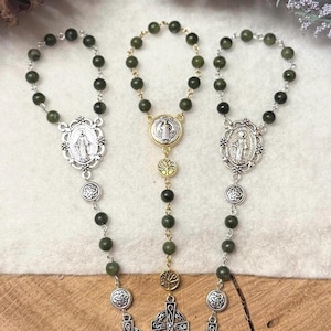Puede incluir: Tres rosarios con cuentas de color verde oscuro y detalles en metal plateado. Dos rosarios tienen medallones ovalados y cruces celtas, mientras que el central presenta una medalla redonda de San Benito, dijes de &aacute;rbol dorados y una cruz celta.