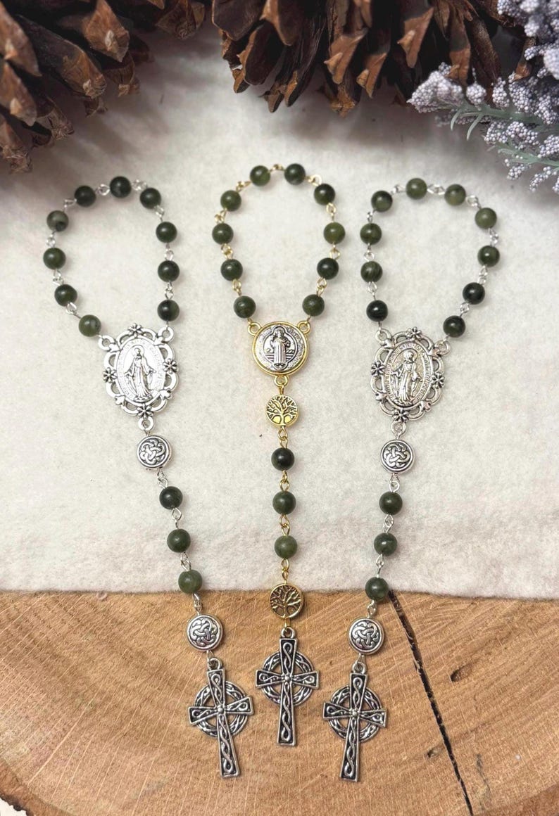 Puede incluir: Tres rosarios con cuentas de color verde oscuro y detalles en metal plateado. Dos rosarios tienen medallones ovalados y cruces celtas, mientras que el central presenta una medalla redonda de San Benito, dijes de &aacute;rbol dorados y una cruz celta.