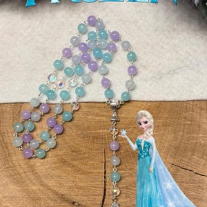 May include: A rosary with light blue, lavender, and clear beads. The rosary has a silver cross pendant and a silver centerpiece. The word "FROZEN" is at the top of the image, and a cartoon image of Elsa is on the right.