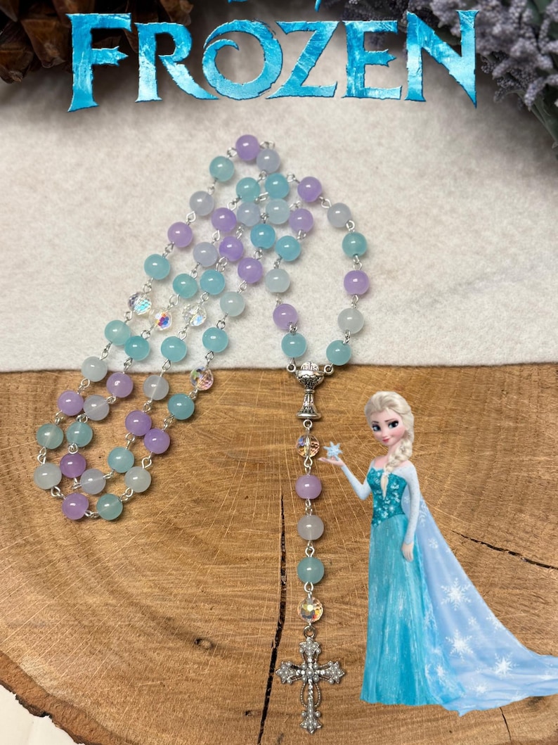 May include: A rosary with light blue, lavender, and clear beads. The rosary has a silver cross pendant and a silver centerpiece. The word "FROZEN" is at the top of the image, and a cartoon image of Elsa is on the right.