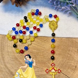 May include: A colorful rosary with yellow, red, blue, and black beads. The rosary features a gold-colored crucifix and a medallion. A cartoon image of Snow White is in the lower left corner of the image.