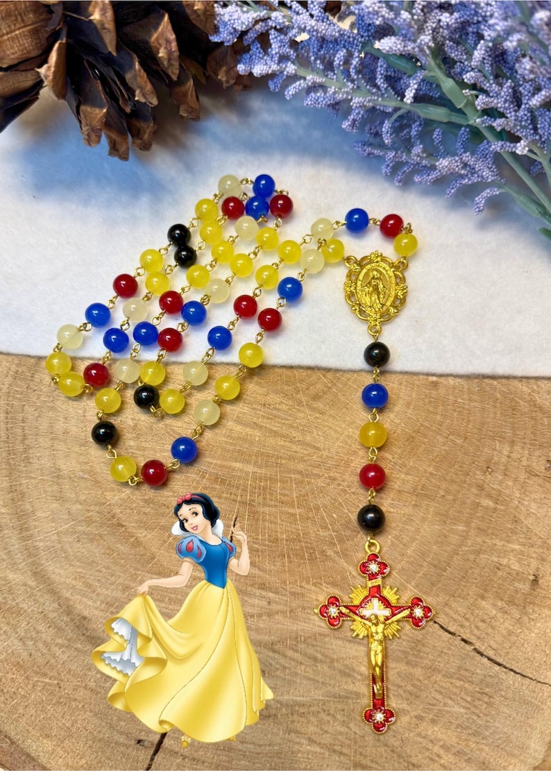 May include: A colorful rosary with yellow, red, blue, and black beads. The rosary features a gold-colored crucifix and a medallion. A cartoon image of Snow White is in the lower left corner of the image.