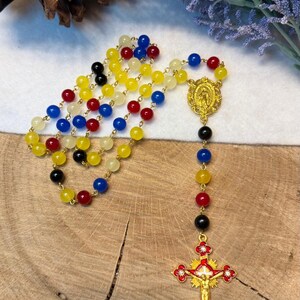May include: A colorful rosary with beads in yellow, red, blue, and black. The rosary features a gold-colored crucifix with a figure of Jesus and a gold-colored centerpiece with a religious image. The beads are strung on a gold chain.