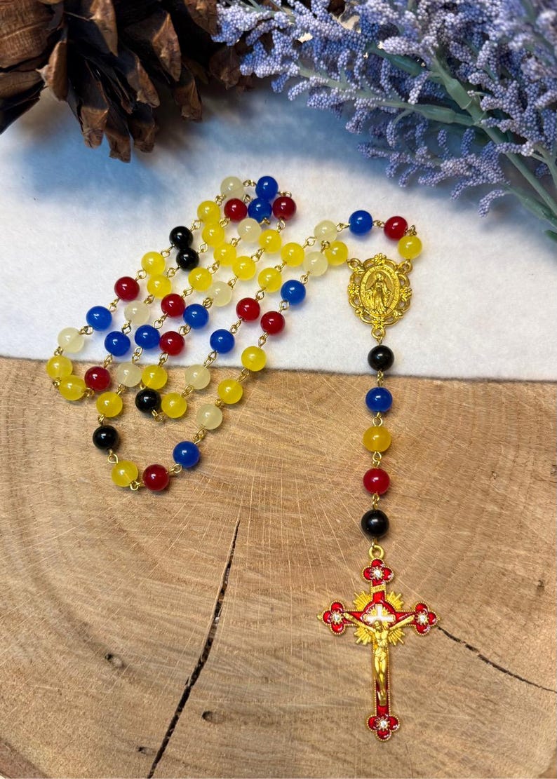 May include: A colorful rosary with beads in yellow, red, blue, and black. The rosary features a gold-colored crucifix with a figure of Jesus and a gold-colored centerpiece with a religious image. The beads are strung on a gold chain.