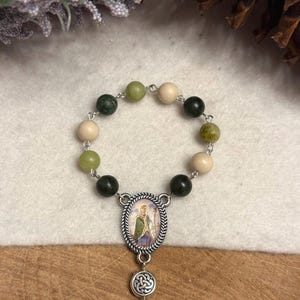 Puede incluir: Un rosario religioso con una disposici&oacute;n circular de cuentas que alternan entre verde oscuro, verde claro y beige. La pieza central es un colgante ovalado de metal plateado que representa una figura con t&uacute;nica verde, seguido de un nudo celta.