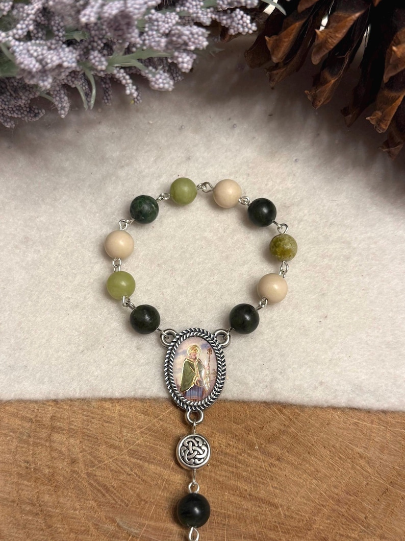 Puede incluir: Un rosario religioso con una disposici&oacute;n circular de cuentas que alternan entre verde oscuro, verde claro y beige. La pieza central es un colgante ovalado de metal plateado que representa una figura con t&uacute;nica verde, seguido de un nudo celta.