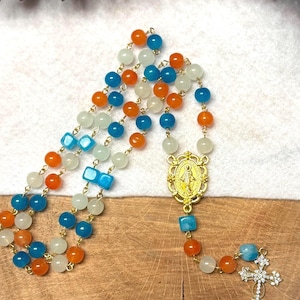 May include: A rosary with alternating orange, blue, and white beads. The rosary features a gold-colored centerpiece with a religious figure and a cross pendant with clear crystal accents. The beads are strung on a gold-colored chain.