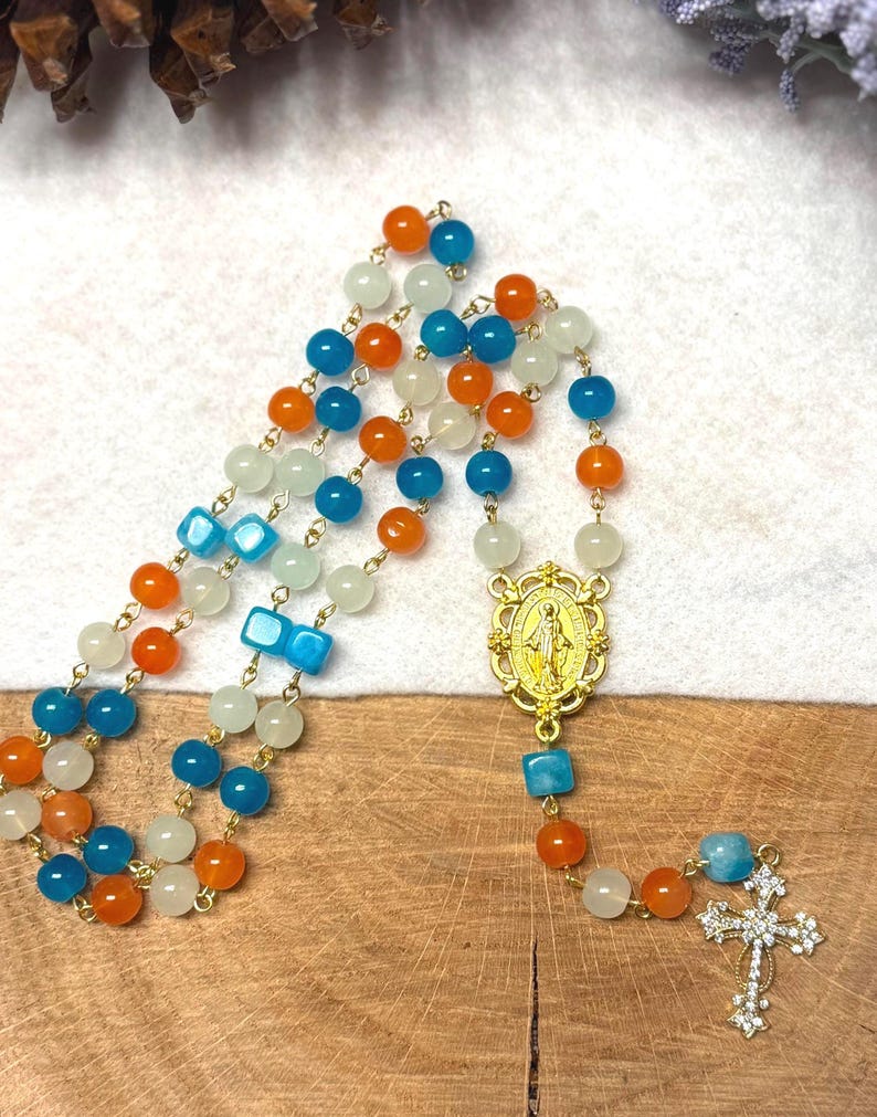 May include: A rosary with alternating orange, blue, and white beads. The rosary features a gold-colored centerpiece with a religious figure and a cross pendant with clear crystal accents. The beads are strung on a gold-colored chain.