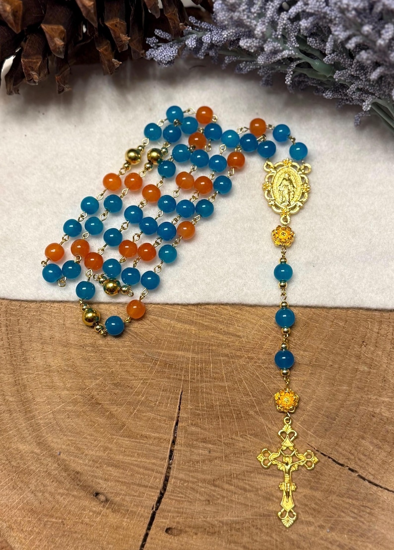 May include: A rosary with alternating blue and orange beads. The rosary features gold-colored metal accents, including a crucifix and a pendant with a religious figure. The beads are strung on a gold chain.