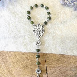 Puede incluir: Un rosario con cuentas de color verde oliva oscuro unidas por eslabones de metal plateado. La pieza incluye una medalla ovalada central con una figura, dos dijes circulares con dise&ntilde;o de &aacute;rbol y un colgante de cruz de estilo celta plateado.