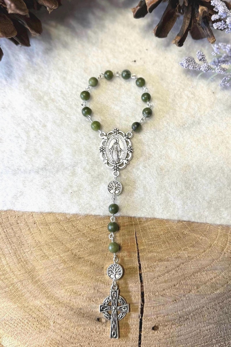 Puede incluir: Un rosario con cuentas de color verde oliva oscuro unidas por eslabones de metal plateado. La pieza incluye una medalla ovalada central con una figura, dos dijes circulares con dise&ntilde;o de &aacute;rbol y un colgante de cruz de estilo celta plateado.