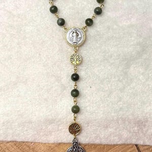 Puede incluir: Un rosario con cuentas de color verde oscuro unidas por eslabones de metal dorado. Incluye una medalla de San Benito plateada, dos dijes circulares dorados con forma de &aacute;rbol y un colgante de cruz celta plateada en la parte inferior.