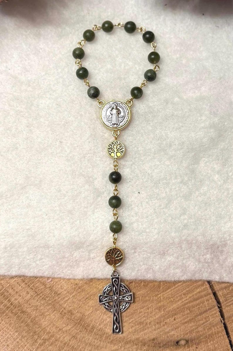 Puede incluir: Un rosario con cuentas de color verde oscuro unidas por eslabones de metal dorado. Incluye una medalla de San Benito plateada, dos dijes circulares dorados con forma de &aacute;rbol y un colgante de cruz celta plateada en la parte inferior.