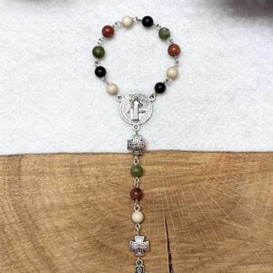 Puede incluir: Un rosario de una d&eacute;cada con un bucle circular de cuentas de piedra en colores negro, crema, verde oliva y marr&oacute;n rojizo, unidas por eslabones de metal plateado. Incluye una medalla central, dos peque&ntilde;as cruces y un crucifijo de estilo celta.