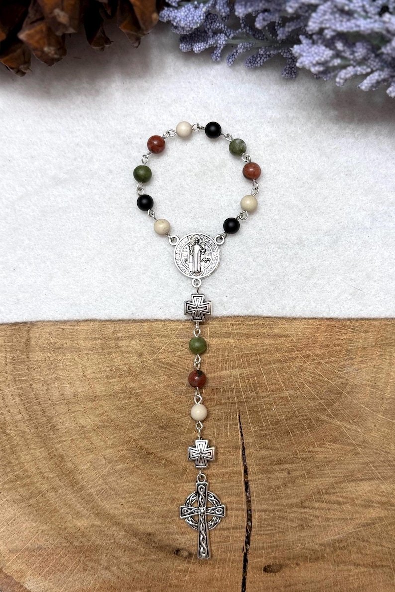 Puede incluir: Un rosario de una d&eacute;cada con un bucle circular de cuentas de piedra en colores negro, crema, verde oliva y marr&oacute;n rojizo, unidas por eslabones de metal plateado. Incluye una medalla central, dos peque&ntilde;as cruces y un crucifijo de estilo celta.