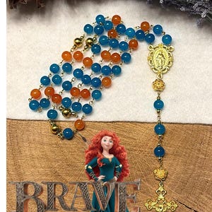 May include: A rosary with blue and orange beads, gold-colored connectors, and a gold-colored crucifix. The rosary is displayed with a cartoon image of a woman and the word "BRAVE".