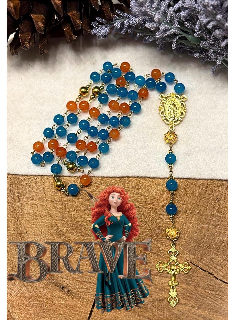 May include: A rosary with blue and orange beads, gold-colored connectors, and a gold-colored crucifix. The rosary is displayed with a cartoon image of a woman and the word "BRAVE".