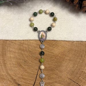 Puede incluir: Un rosario religioso que presenta un bucle circular de cuentas alternadas en verde oscuro, verde claro y color crema. Incluye una medalla ovalada central con la imagen de un santo, dos espaciadores de nudo celta plateados y un crucifijo.