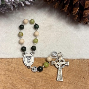 Puede incluir: Un rosario con cuentas alternas de color verde oscuro, verde claro y crema conectadas por eslabones de metal plateado. Incluye una medalla ovalada central con el texto "Pray for us" y una cruz celta plateada con detalles de nudos.