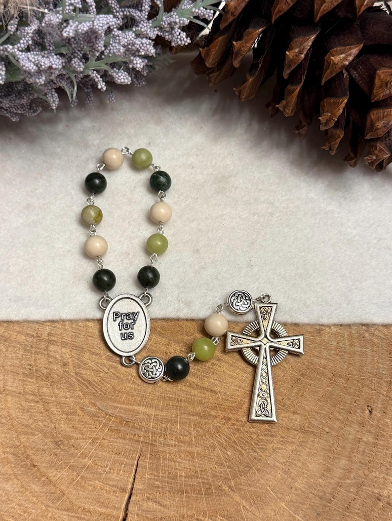Puede incluir: Un rosario con cuentas alternas de color verde oscuro, verde claro y crema conectadas por eslabones de metal plateado. Incluye una medalla ovalada central con el texto "Pray for us" y una cruz celta plateada con detalles de nudos.