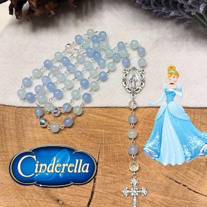 May include: A rosary with light blue and clear beads, a silver cross pendant, and a Cinderella graphic. The rosary features a silver centerpiece with a religious figure. The word "Cinderella" is displayed in a blue and gold graphic.