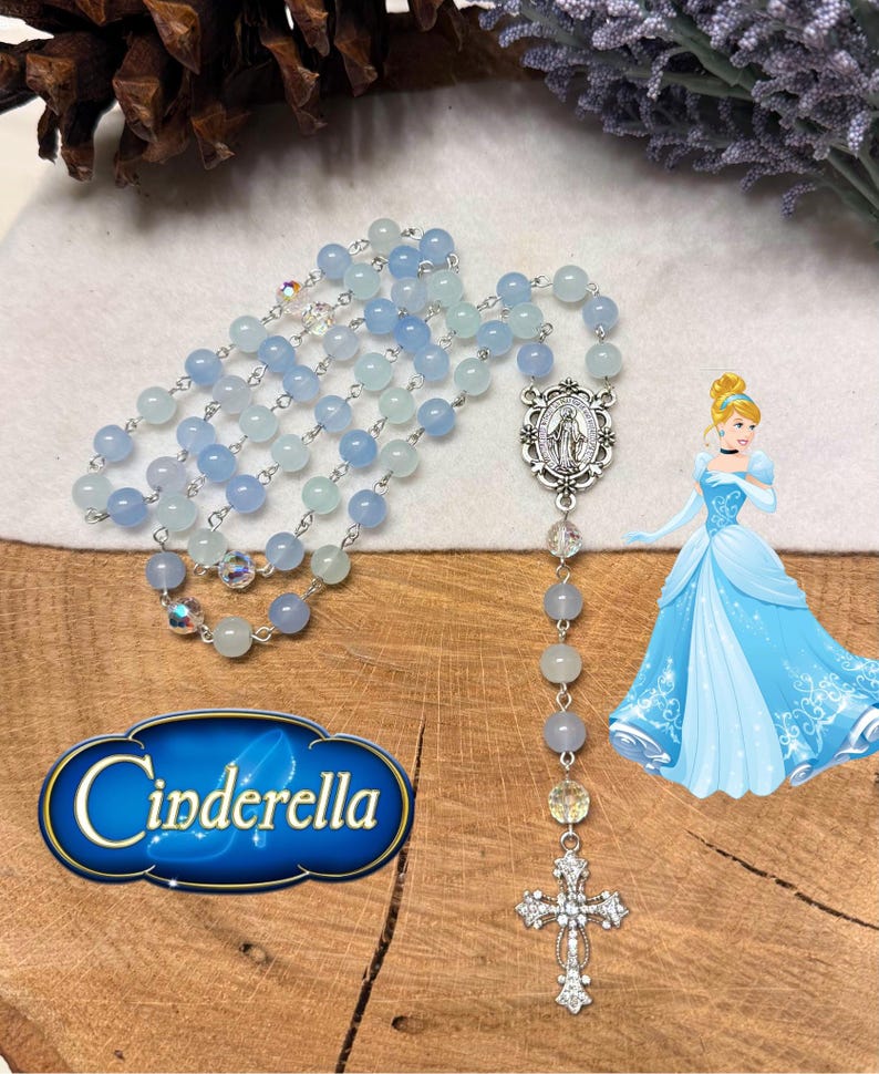 May include: A rosary with light blue and clear beads, a silver cross pendant, and a Cinderella graphic. The rosary features a silver centerpiece with a religious figure. The word "Cinderella" is displayed in a blue and gold graphic.