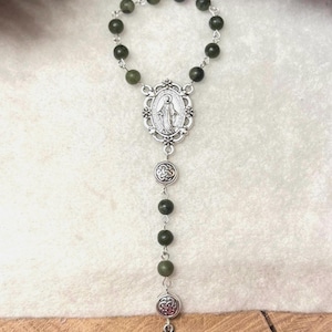 Puede incluir: Un rosario con cuentas de color verde oscuro unidas por eslabones de metal plateado. La pieza central es una medalla ovalada que representa una figura, seguida de dos dijes circulares con patrones de nudos y un colgante de cruz celta plateada.