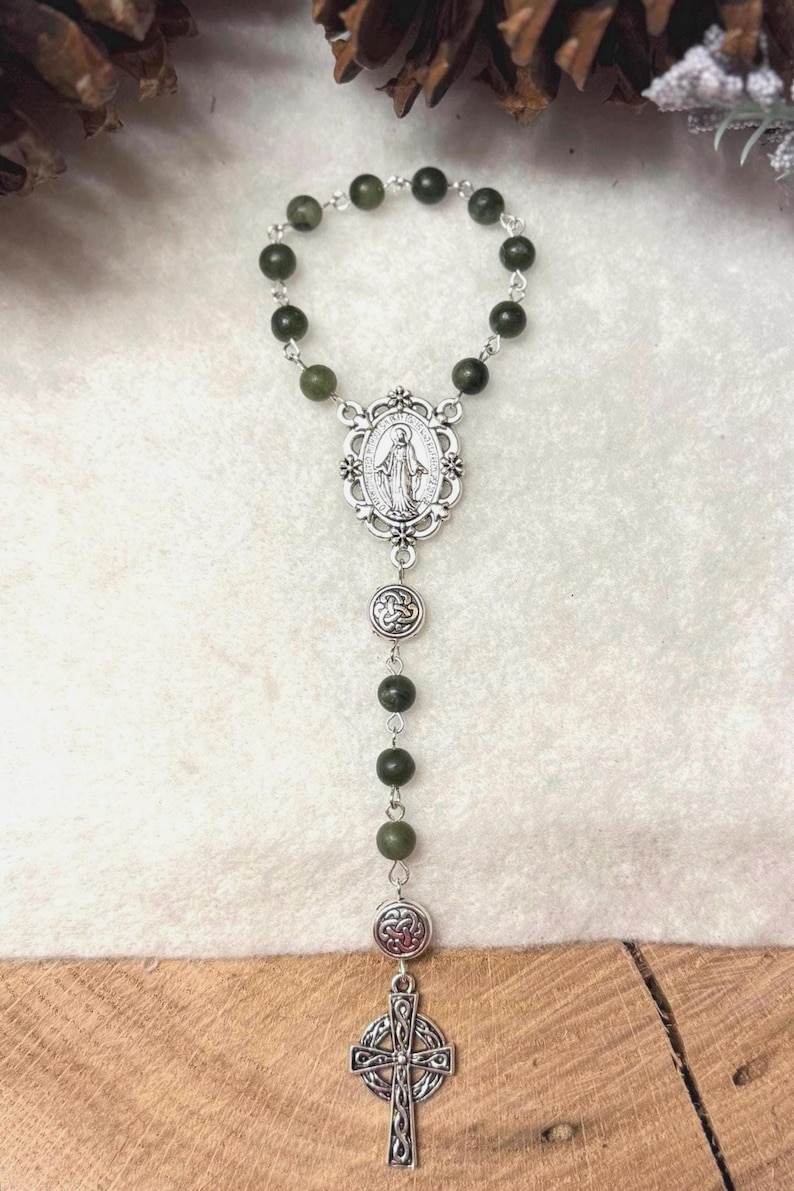 Puede incluir: Un rosario con cuentas de color verde oscuro unidas por eslabones de metal plateado. La pieza central es una medalla ovalada que representa una figura, seguida de dos dijes circulares con patrones de nudos y un colgante de cruz celta plateada.