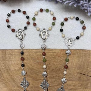 Puede incluir: Tres pulseras de rosario con eslabones de metal plateado, medallones religiosos circulares con figuras y peque&ntilde;os dijes de cruz. Las pulseras est&aacute;n compuestas por cuentas redondas en colores negro, marr&oacute;n, verde y crema.