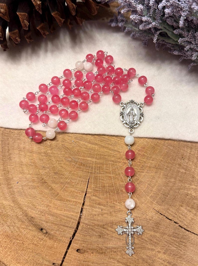 May include: A rosary with pink and white beads. The beads are strung on a silver chain. The rosary features a silver cross and a silver pendant with an image of the Virgin Mary.