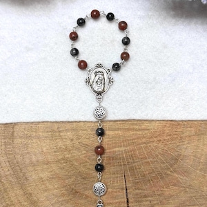 Puede incluir: Un rosario con cuentas alternas de color rojo oscuro y negro unidas por eslabones de metal plateado. La pieza incluye una medalla ovalada central con una figura, dos dijes circulares con dise&ntilde;os de nudos intrincados y un colgante de cruz celta.