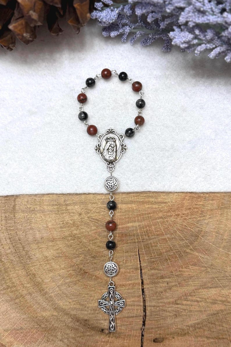 Puede incluir: Un rosario con cuentas alternas de color rojo oscuro y negro unidas por eslabones de metal plateado. La pieza incluye una medalla ovalada central con una figura, dos dijes circulares con dise&ntilde;os de nudos intrincados y un colgante de cruz celta.