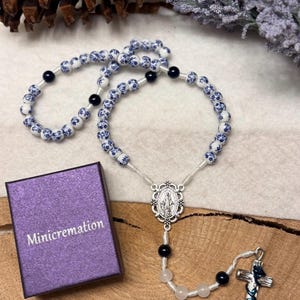 May include: A rosary with blue and white floral beads, black beads, and a silver cross. The rosary is next to a purple box with the word "Minicremation" printed on it. The rosary is on a wooden surface.