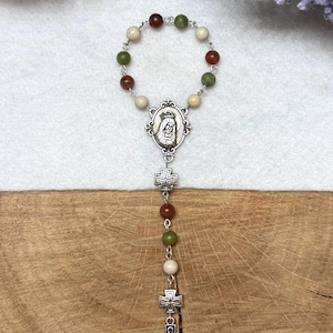 Puede incluir: Un rosario de una decena con un bucle circular de cuentas alternas rojas, verdes y crema unidas por eslabones de metal plateado. Una medalla plateada que representa una figura con corona se sit&uacute;a sobre dos peque&ntilde;as cruces y una cruz celta.