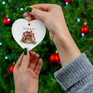 Peut inclure: Ornement en céramique blanche en forme de cœur avec le mot "Bookoholic" au-dessus d'une pile de livres liés avec des rubans rouges et verts. L'ornement est tenu par une personne devant un sapin de Noël.
