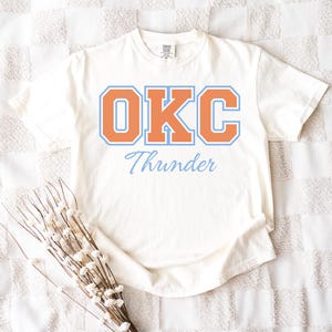 Op de afbeelding: Crèmekleurig T-shirt met de tekst "OKC Thunder" in oranje en lichtblauw. Het shirt heeft een klassiek, college-achtig ontwerp, ideaal voor fans van het basketbalteam Oklahoma City Thunder.