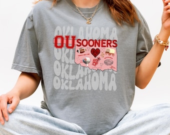 May include: Gray t-shirt featuring the text "OKLAHOMA" in a repeating pattern. The shirt has a graphic of the state of Oklahoma with "OU SOONERS" and various icons. The graphic includes a football, heart, and covered wagon.