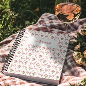 Könnte beinhalten: Ein Spiralnotizbuch mit weißem Einband, verziert mit einem Muster aus rosa Schleifen und floralen Elementen. Das Wort "Notebook" ist in Schreibschrift geschrieben. Ein Glas Roséwein steht auf einer karierten Decke, was eine Picknick-Szene suggeriert.
