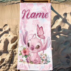 Pode incluir: Toalha de praia rosa com um personagem de desenho animado, uma borboleta e flores. A toalha tem a palavra "Name" em uma fonte cursiva. O fundo tem um padr&atilde;o floral. A toalha &eacute; exibida em uma cadeira de praia de madeira em uma praia arenosa.