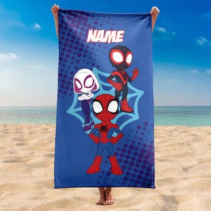 May include: Blue beach towel featuring cartoon Spider-Man characters. The towel has the word "NAME" at the top. The characters are in a web design with a dotted background. The towel is held up on a beach.