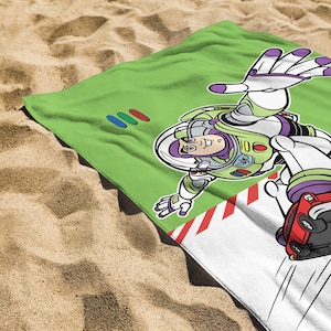 May include: A green beach towel featuring a cartoon illustration of Buzz Lightyear in a dynamic pose. The design includes a red race car and colorful accents. The towel is on a sandy surface.