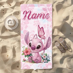Pode incluir: Toalha de praia rosa com um personagem de desenho animado, uma borboleta e flores. A toalha tem a palavra "Name" em escrita rosa. Um chap&eacute;u branco e sand&aacute;lias est&atilde;o na areia.
