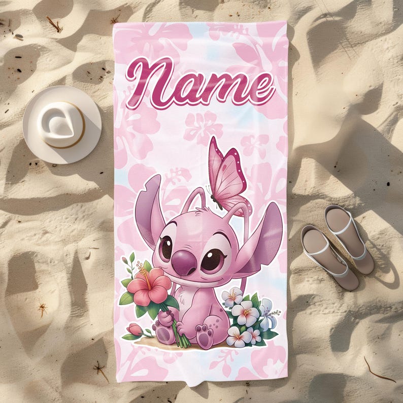 Pode incluir: Toalha de praia rosa com um personagem de desenho animado, uma borboleta e flores. A toalha tem a palavra "Name" em escrita rosa. Um chap&eacute;u branco e sand&aacute;lias est&atilde;o na areia.