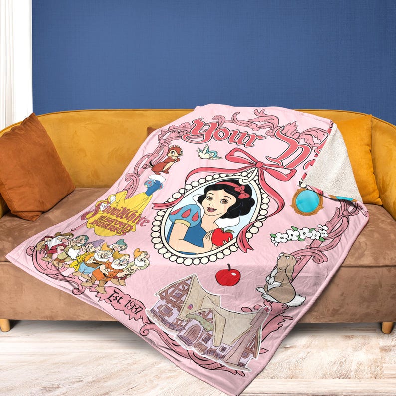 May include: Pink throw blanket featuring Snow White and the Seven Dwarfs. The blanket has a framed image of Snow White holding an apple, with other characters and the text "Est. 1937". The blanket is draped over a sofa.