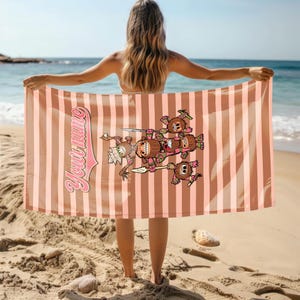 May include: A pink and brown striped beach towel with a cartoon illustration of a group of brown and pink characters holding spears. The text "Your Name" is written in a pink and white font on the towel.