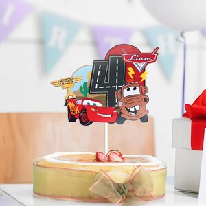 May include: A cake topper featuring Lightning McQueen and Mater from the Cars movie, with a large number 4 and the name "Liam". The topper is placed on a cake decorated with strawberries and a ribbon.
