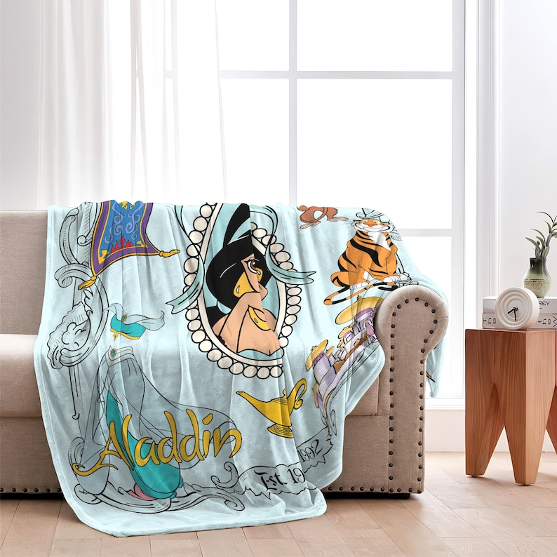 May include: A light blue throw blanket featuring illustrations from the Disney movie Aladdin. The blanket displays Jasmine in a pearl frame, a magic carpet, a tiger, and the word "Aladdin" in gold script. The blanket is draped over a beige sofa.
