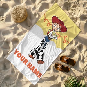 May include: A white beach towel featuring a cartoon of a cowgirl with a red hat and cow print pants. The towel has the text "YOUR NAME" in orange. A straw hat and brown sandals are also visible.