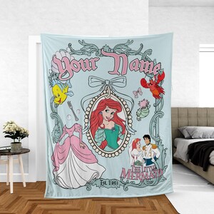 May include: Light blue blanket featuring a framed portrait of a red-haired woman in a green dress, surrounded by cartoon characters and the text "Your Name" and "The Little Mermaid". The blanket has a decorative border and the text "Est. 1989".